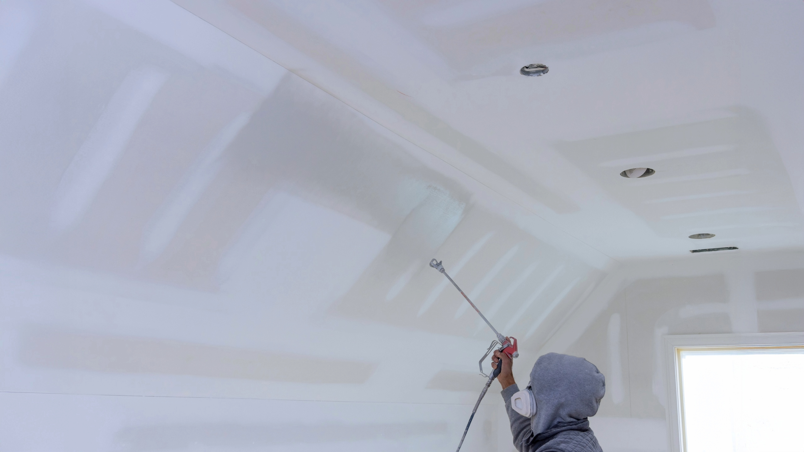 Professional painter using an airless sprayer during an interior house painting project to coat newly finished drywall ceilings in a residential home.
