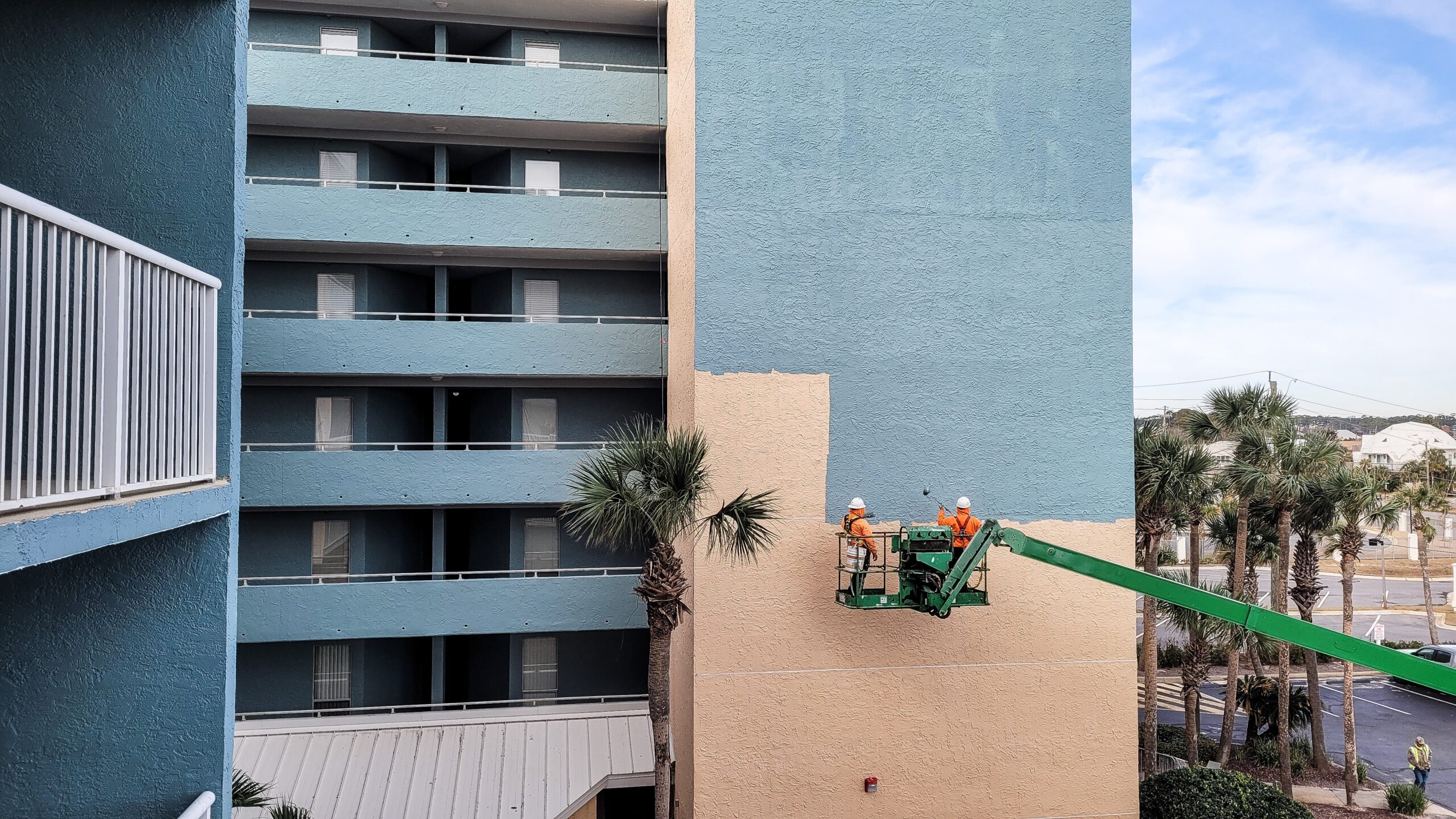 Two commercial painters in orange safety vests and white helmets are elevated on a lift, painting the side of a tall hotel building from tan to blue. The building has several floors of balconies, palm trees in the foreground, and a clear sky overhead.