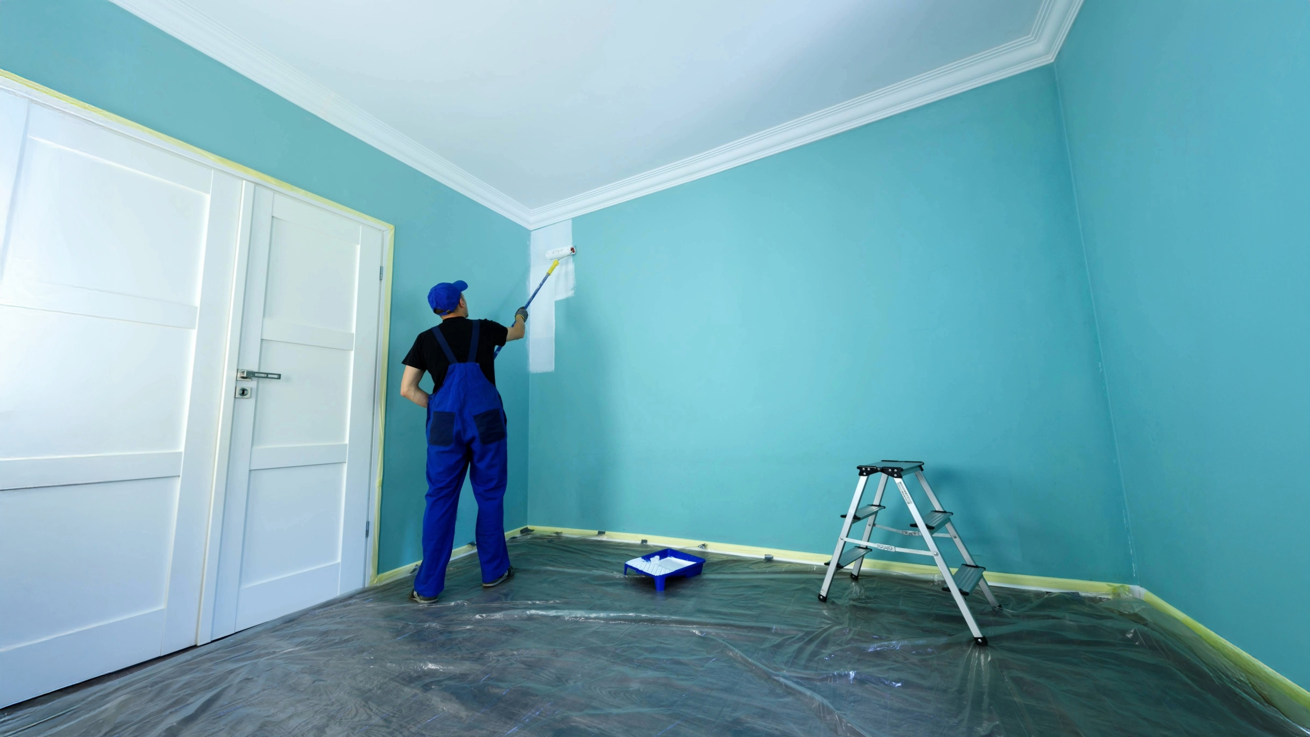 Professional painter in blue overalls applying white paint over a turquoise interior wall using a roller, with a step ladder and painting tray nearby in a prepared room with plastic floor covering.