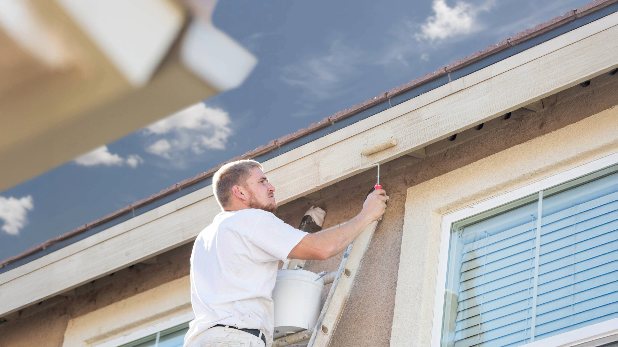 Professional painter using roller brush to paint exterior house siding while standing on ladder under clear sky.